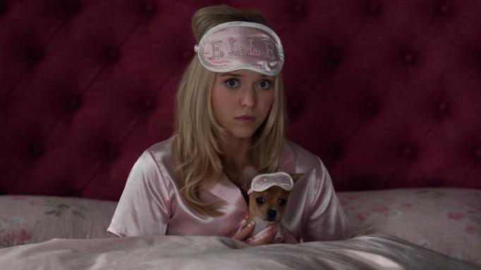A woman with long blonde hair sits up in bed, with pink bedsheets, pink satin PJs and a pink eye mask on her forehead that has 