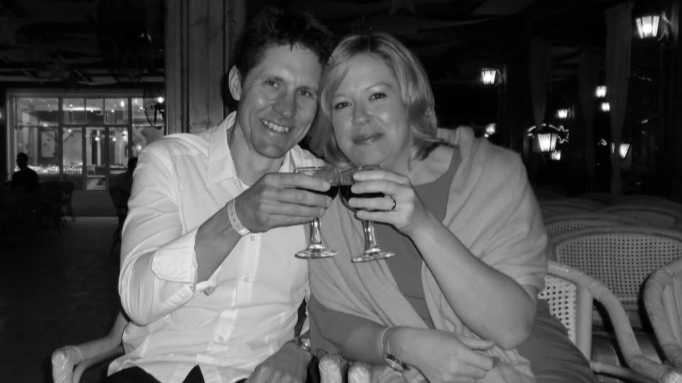 A man and a woman are making a toast with their wine glasses. They are pictured in a black and white photo. The man is wearing a white shirt and has short hair. The woman is wearing a dress with a shawl over her shoulders.
