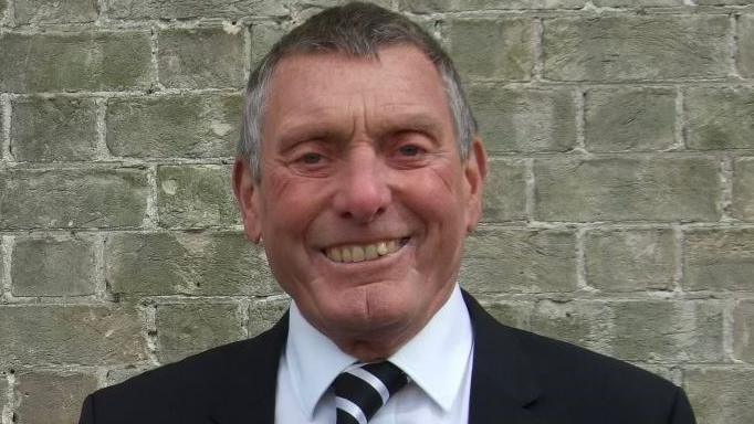 Dennis has short grey-brown hair and is wearing a black suit jacket over a white shirt and a monochrome striped tie