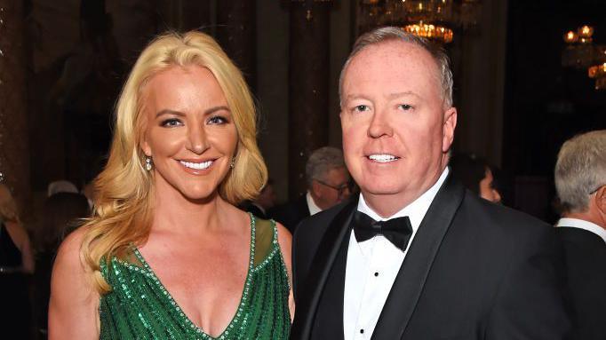 Michelle Mone with blonde hair, shining white teeth, and an emerald green dress stands beside Doug Barrownman, balding with grey hair, in a black tux, bow tie pose for a photo at a posh function
