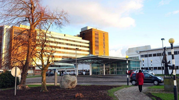 Sbows a hospital with trees in the front, some cars, a statue and a woman in a red overcoat with black trousers 