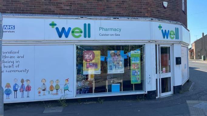 The Well Pharmacy on the junction of High Street and Beach Road in Caister. The building dates from the 1950s and has Crittall windows on the upper storey, and a façade with large windows covered in a vinyl depicting families in a child-like drawing. Houses on High Street can be seen on the right of the image.