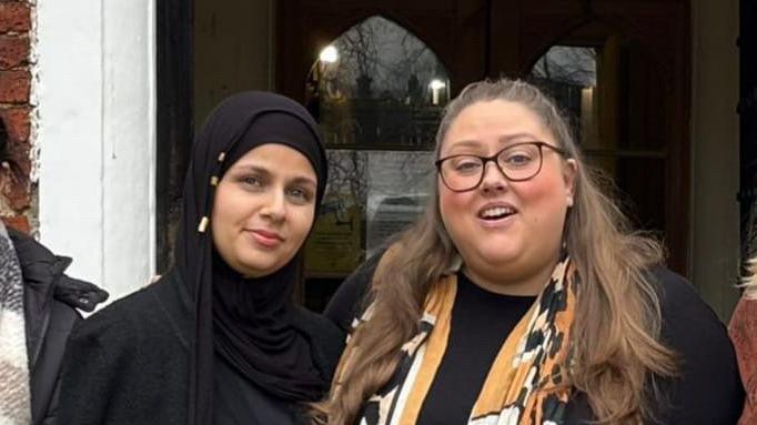 Jwan Mustafa and Emma Parson stand side by side smiling at the camera. Ms Mustafa wears a black head scarf while Ms Parson is wearing a black top and a brown scarf around her neck.