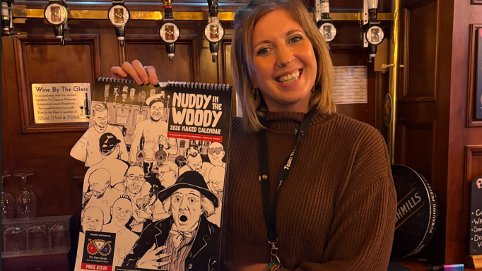 A woman is stood behind a pub bar, holding a calendar with cartoon drawings of men on the front. The calendar is called Nuddy in the Woody