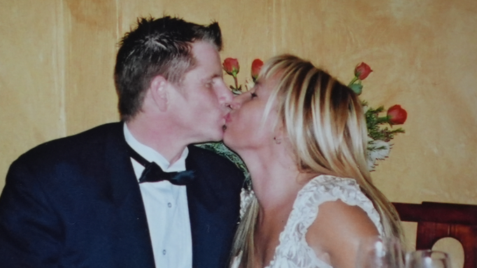 A view in profile of a man in a tuxedo and and black bow tie and woman in a white dress giving each other a peck on the lips 