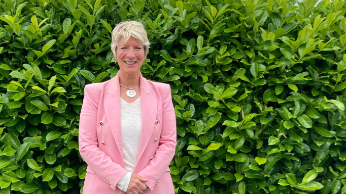 Woman with short blonde hair wearing a pink jacket and white top with a green bush in the background