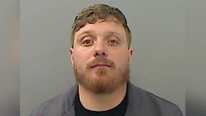 A mugshot of Jack Walker who has light brown short hair which sweeps to the side. He also has a beard.