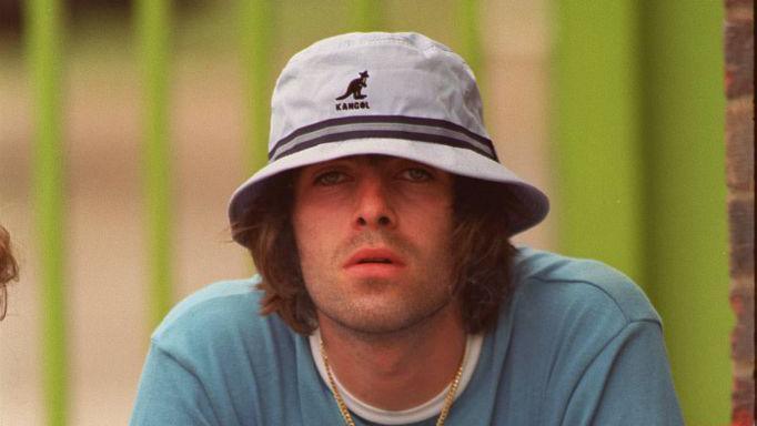 Liam at the Music Industry Soccer Six Tournament at Mile End Stadium, East London in May 1996. He is wearing a blue t-shirt, gold chain necklace, and a white and blue striped Kangol bucket hat.