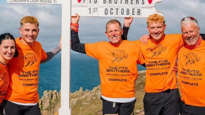 Four men and one woman in orange t-shirts printed with a cartoon logo of a brain with legs, saying The FTD Brothers and Complete, stand with their arms raised and smiling next to a white signpost. They are on a clifftop, with the sea visible in the background. 