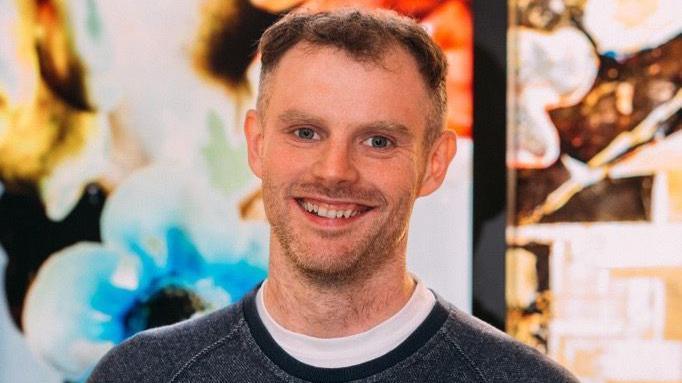 A man with dark blond hair stands in front of a colourful background, pictured from the head and shoulders up. He wears a white T-shirt and black jumper.
