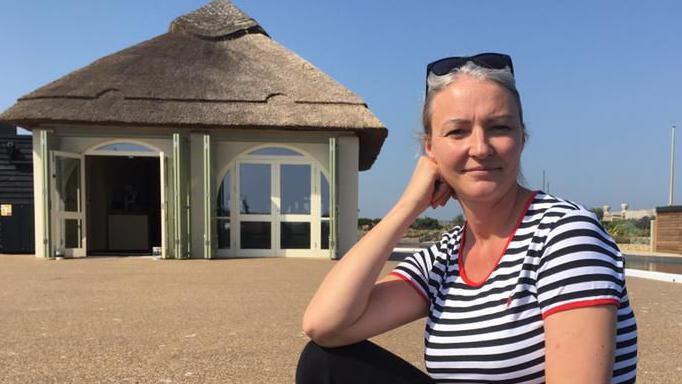 Emma Ratzer, sitting on the deck by the boating lake in Great Yarmouth, wearing black trousers, a black and white striped tee shirt with red trim, and she has sunglasses up in her greying, tied back hair. Behind her is the boating lake cafe.