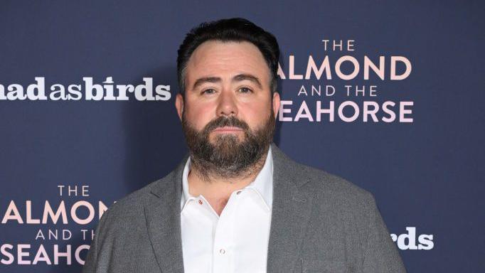 Celyn Jones wearing a grey blazer and a white shirt. He is standing in front of a dark blue screen. He has short dark hair and a short beard. 