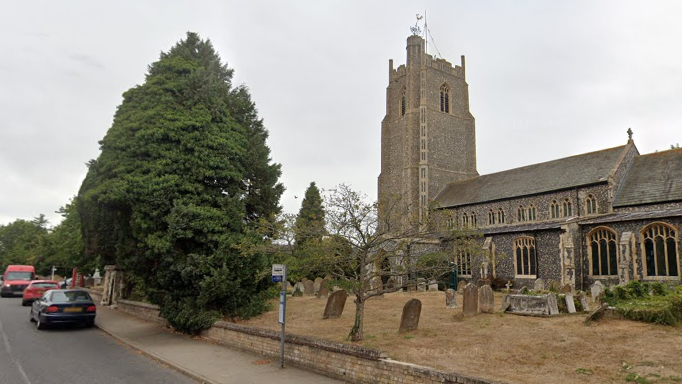 A general view of Church Street in Stradbroke. To the right is the large, church, made out of grey and brown stones, and its spire. There are various graves dotted throughout the cemetery in front of the church. To the left is the road, with three cars parked next to the kerb.