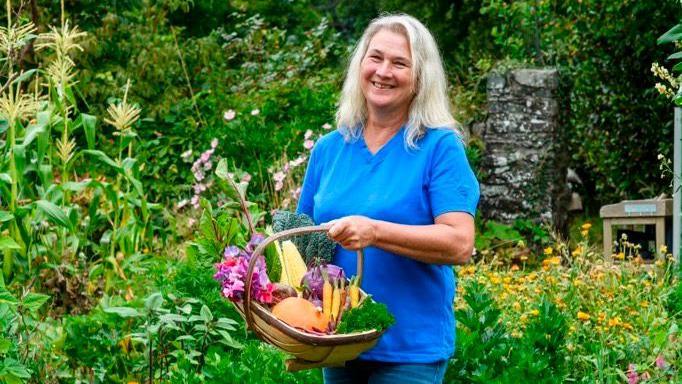 Stephanie Hafferty standing in her garden holding a wicker basket of homegrown vegetables.  She is wearing a blue top and jeans, and has long white hair.  