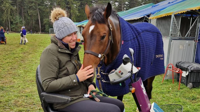 Caroline March in a green waxed jacket and grey bobble hat, who is sitting in a mobility scooter while stroking the nose of chestnut coloured horse. The horse is wearing a blue coat 