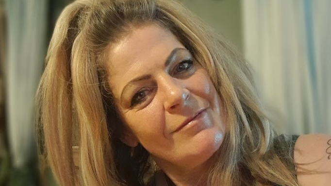 Jay has long blonde hair and is wearing a green top. Her head is slightly tilted to the side and she is smiling at the camera. There is a blurred background of some white curtains.