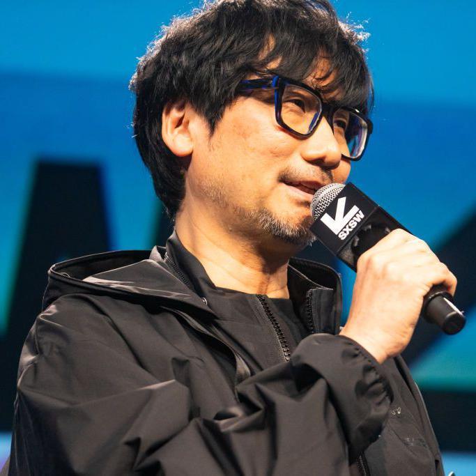 Hideo Kojima. He has black hair in a short cut and wears large black glasses. He is smiling while he talks at a conference. He is wearing a trim black jacket and black t-shirt.