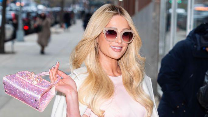 A woman with long blonde hair smiles at the camera while wearing pink sunglasses. She's holding a small sparkly pink handbag