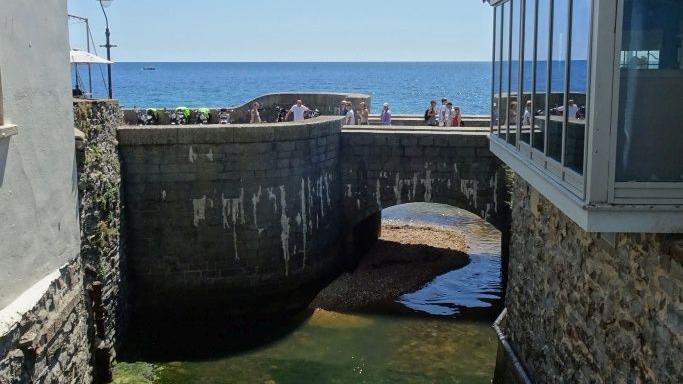 Pedestrians walk over a stone bridge crossing the river Lim where it meets the sea. The river is shallow with high stone walls either side with buildings on top. It's a sunny day and the sea beyond the bridge is calm and bright blue.
