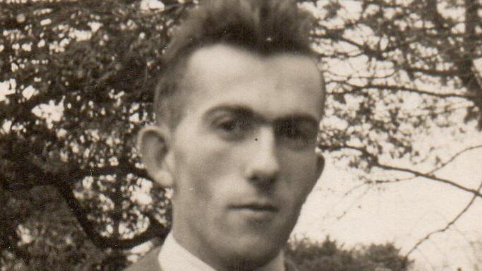 Black and white image of thin young man with short dark hair and sculpted cheekbones, wearing a dark overcoat, shirt and tie, standing in front of a tree.