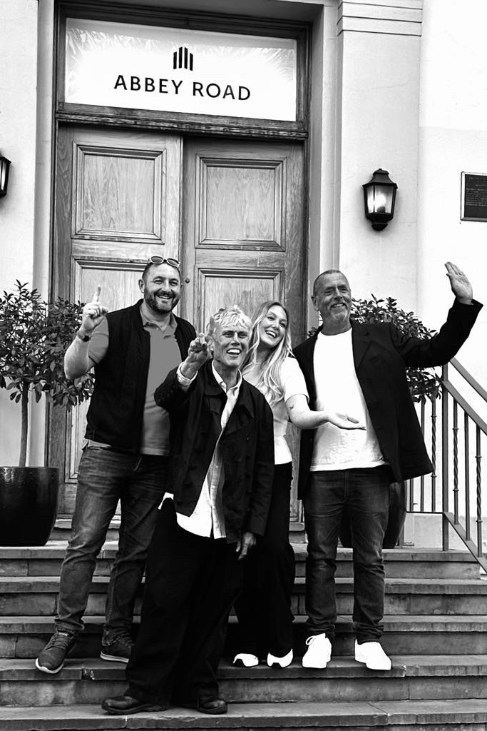A black and white photo of three men and Emily Fern smile at the camera as they stand on steps leading up to a large wooden door with bay trees pots either side of it. The sign above the door reads ABBEY ROAD. One of the men is Bez and another is Phil Hartnoll.