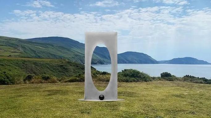 A steel modern art sculpture, which is a rectangle with an oval shaped hole, standing on some grass in front of a scenic view overlooking the sea, with coastal hills stretching into the background. 