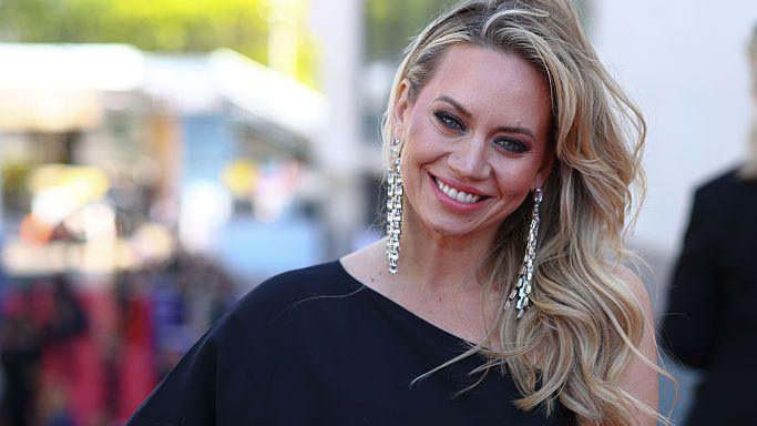 Kimberley Wyatt posed in a one shoulder black dress at a red carpet event. her long blonde hair is curled and swept over one shoulder and she is wearing long silver earrings and smiling.