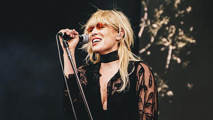 A blonde woman, wearing round red sunglasses, sings into a microphone and closes her eyes.