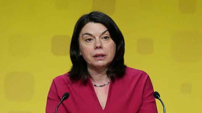 Sarah Olney wearing a purple dress and standing behind a lectern as a Liberal Democrat conference