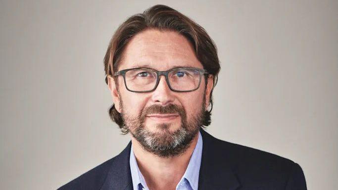 Stuart Simpson wearing a blue collared shirt, a black blazer, and black rectangular framed glasses. He has brown hair cut just below his ears and a full beard with some grey patches in it. He is smiling at the camera standing against a beige background.