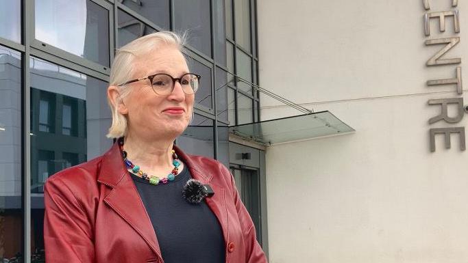 Sally Basker - A woman wearing glasses and a red leather jacket , standing in front of the Science Park centre office block.