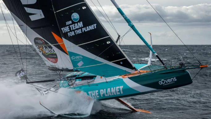 A racing yacht with a white and blue hull and black and white sails going fast with spray behind.