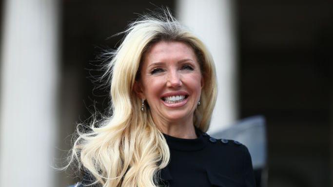 A middle-aged woman with bright blonde hair. She is wearing a black top.