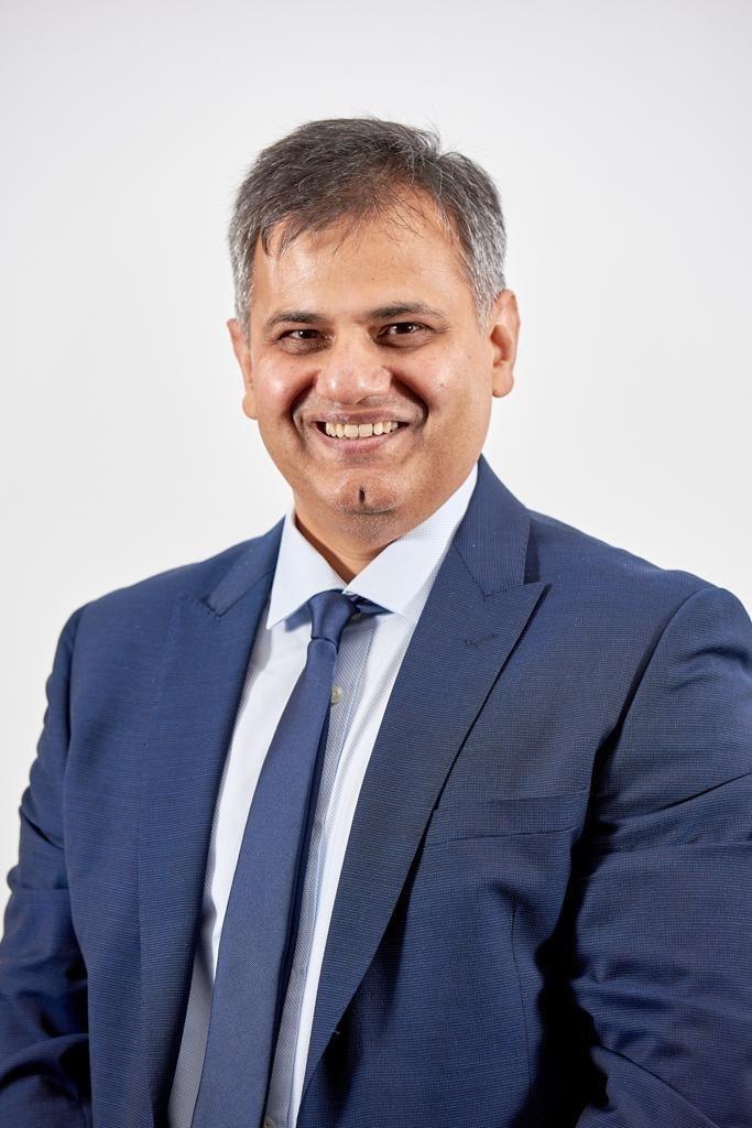 A man wearing a dark blue suit jacket and tie smiles 