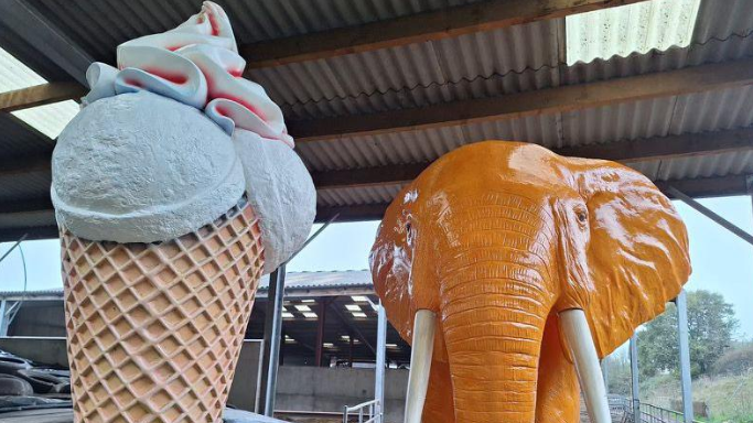 A giant ice cream cone statue next to a large orange elephant statue stood in a barn on a Devon farm