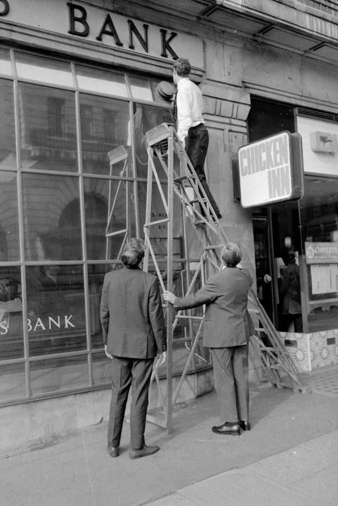 A policeman up a ladder looking at an alarm on the outside of the bank