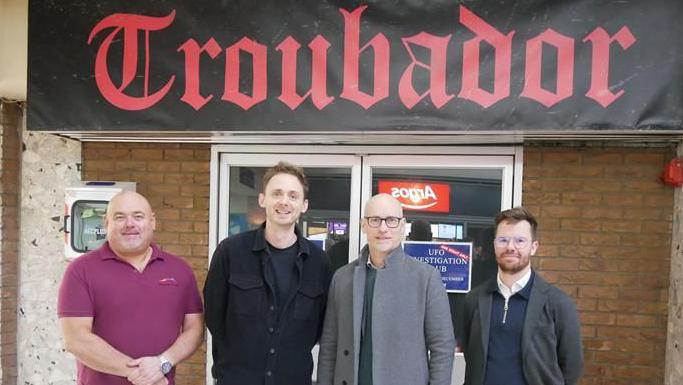 Centre manager Chris Morgan (far left) with photographer Roo Lewis, local MP Stephen Kinnock and James Taylor stand outside double doors with a black banner above with red, gothic style writing with the word 'Troubador' on, recreating the old sign of the club