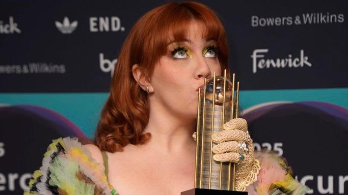 CMAT kissing a gold trophy. She has dark ginger hair with a fringe and is looking up. She is wearing yellow eyeshadow, yellow fishnet gloves and a multi-coloured top. She is standing in front of a Mercury Prize board which has the names of the awards' sponsors on it.