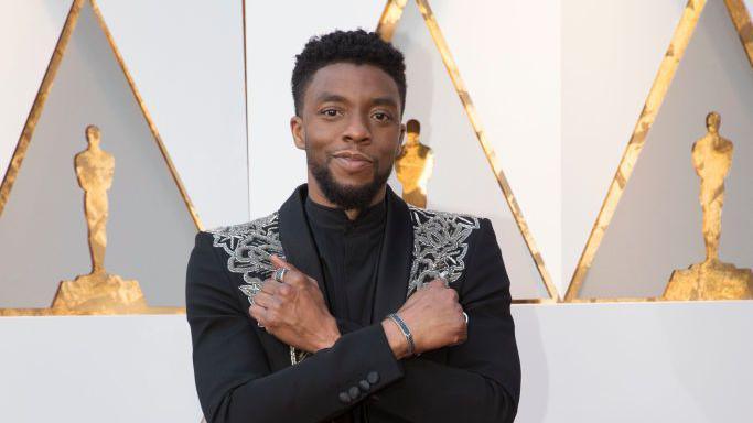 Chadwick Boseman wears a black blazer with silver embellishment. His arms are crossed over his chest in an 'X' shape, which is the Wakanda salute. He is standing in front of a white background with gold Oscar silhouettes.