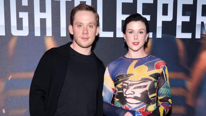 Joe Cole stands to the left of Alexandra Roach. Joe is wearing a black jacket and top while Alexandra wears a multicoloured dress. They both stand behind a poster for the programme with the word 'Nightsleeper' 