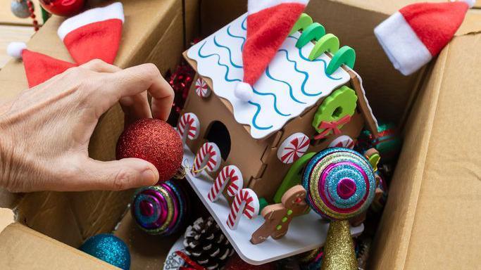 person putting Christmas decorations into a cardboard box - there is a red bauble, a gingerbread house and small Santa hats.