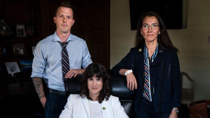 Jared Golden and Mary Peltola stand to the left and right flank respectively of a seated Marie Gluesenkamp Perez as all three look into the camera for a group portrait