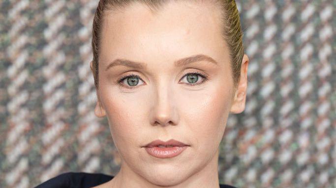 A glamorous red carpet shot of actress Lauren Lyle is a close-up of her face, with a tweed-effect on the background behind her.
