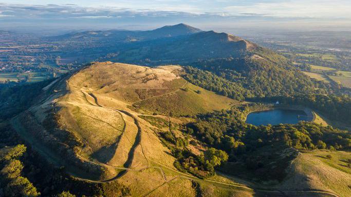 The Malvern Hills with the Iron age hill fort in the foreground. The sun is shining over the landscape in the aerial image.