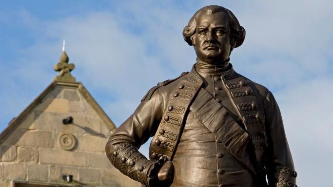 A large bronze coloured statue of a man wearing military clothing, with one hand on his hip.