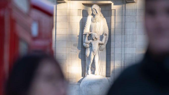 The statue in sunlight with the blurred images of passers-by in the foreground. Red telephone boxes can be seen out of focus on the left of the picture