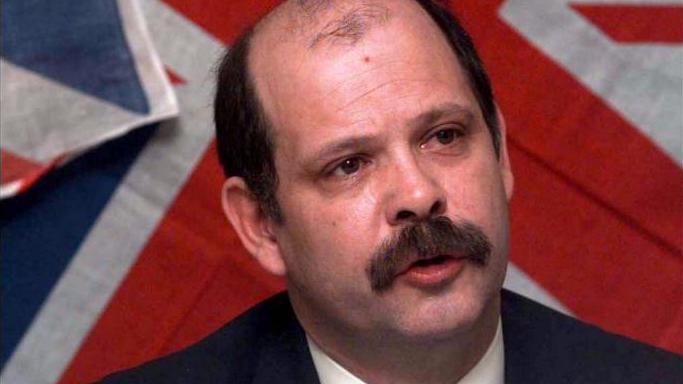 A man with brown hair and moustache in font of a union jack background