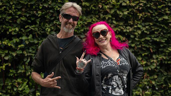 A man and a woman stand in front of a hedge. The man is wearing all black. The woman has bright pink hair, a black leather jacket, and a Black Sabbath T-shirt. They both wear sunglasses.