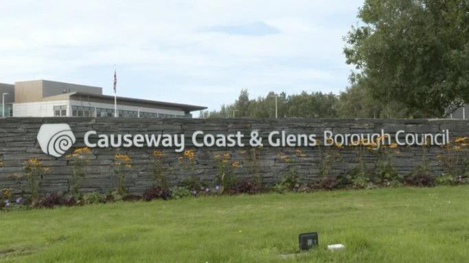 A picture of the offices of Causeway Coast and Glens Council.
There is a picture of a sign saying ' Causeway Coast & Glens Borough Council' on a bricked wall with a grassy patch in front of it. The office building can be seen in the background.