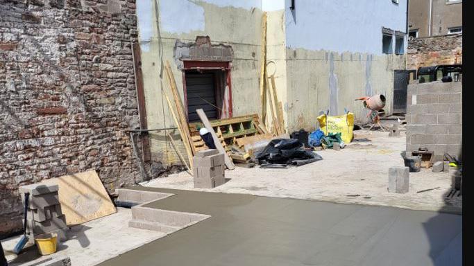 The side of a building which has been stripped down by demolition. There is a newly-laid cement floor with building equipment, including a cement mixer, and materials to the side of it.